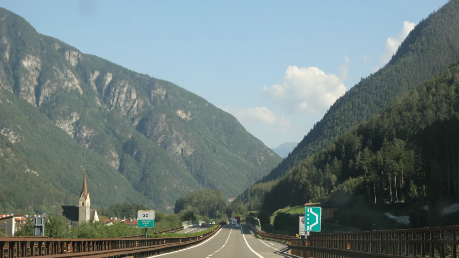 Brennerautobahn A22 bei Mittewald in Südtirol mit zwei Fahrstreifen zwischen bewaldeten Berghängen, links eine Kirche und das Kilometerschild Bolzano Bozen 47, rechts ein grünes Hinweisschild