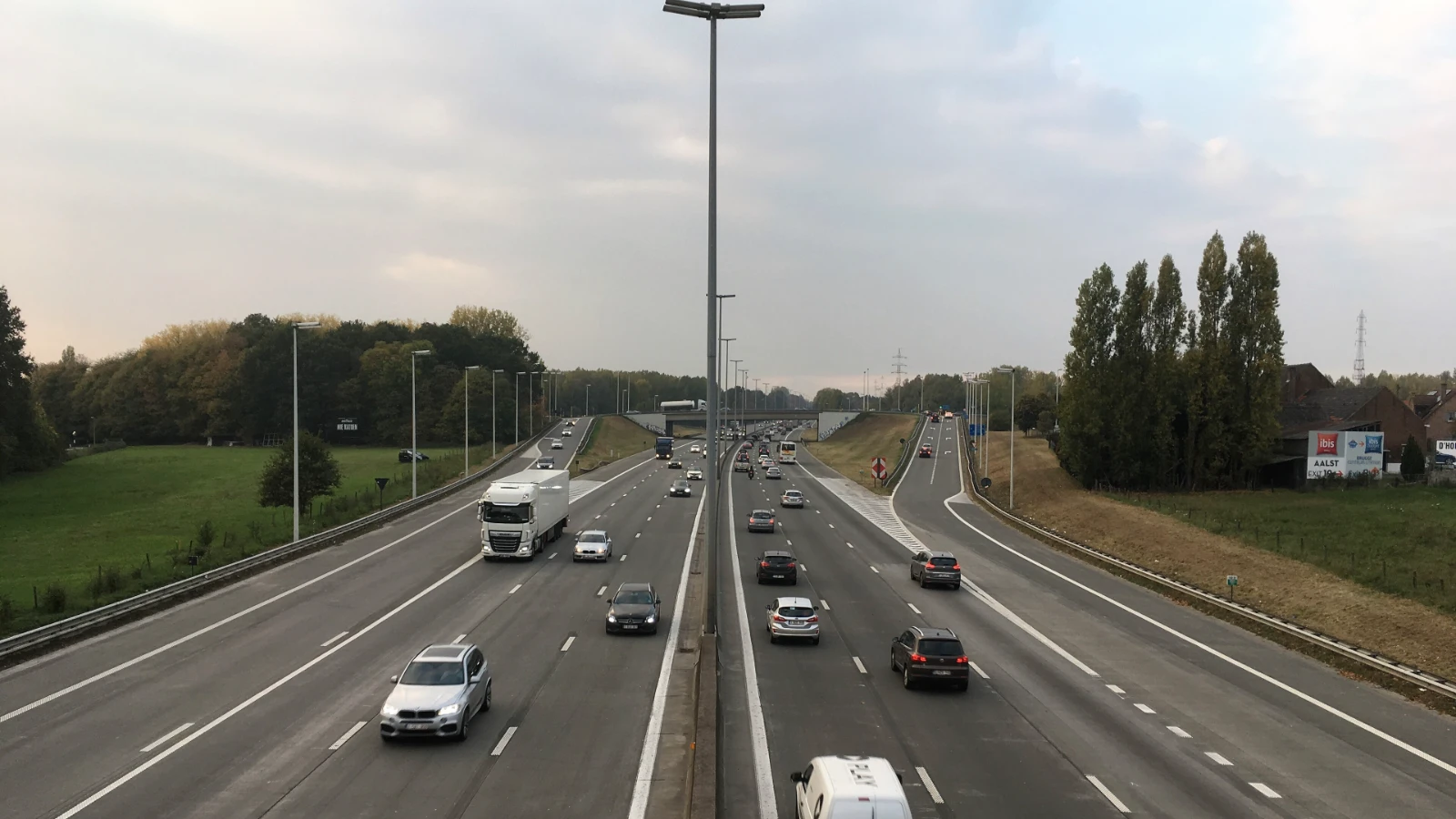 Mehrspurige Autobahn E40 bei Affligem von einer Brücke aus gesehen, Autos, ein Lkw und ein Transporter in beiden Fahrtrichtungen, rechts eine Ausfahrt und eine Reihe Pappeln