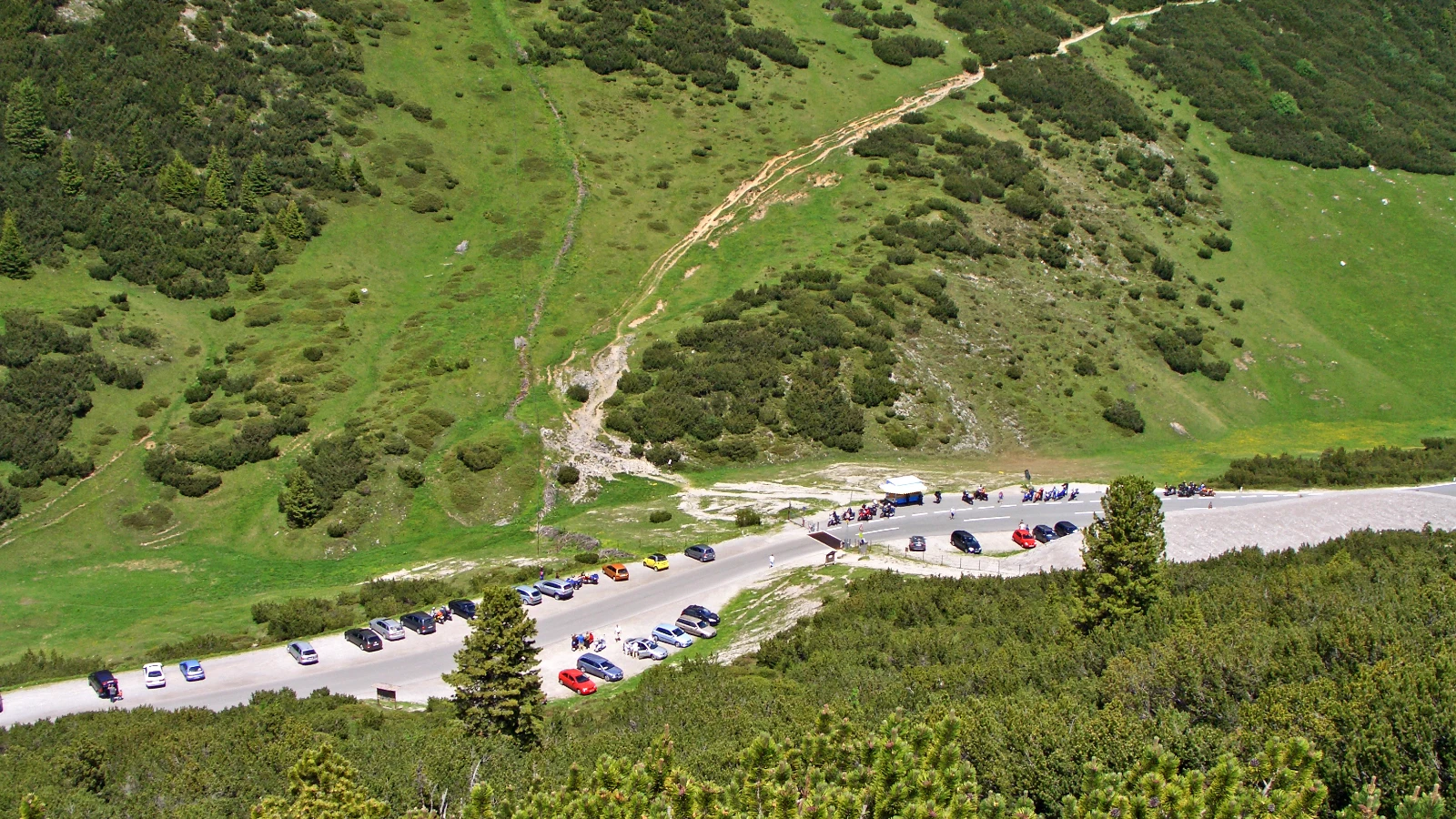 Blick von oben auf die Passhöhe des Hahntennjochs mit geparkten Autos am Straßenrand und einer Reihe abgestellter Motorräder vor einem Kiosk, dahinter grüne Berghänge mit Latschen
