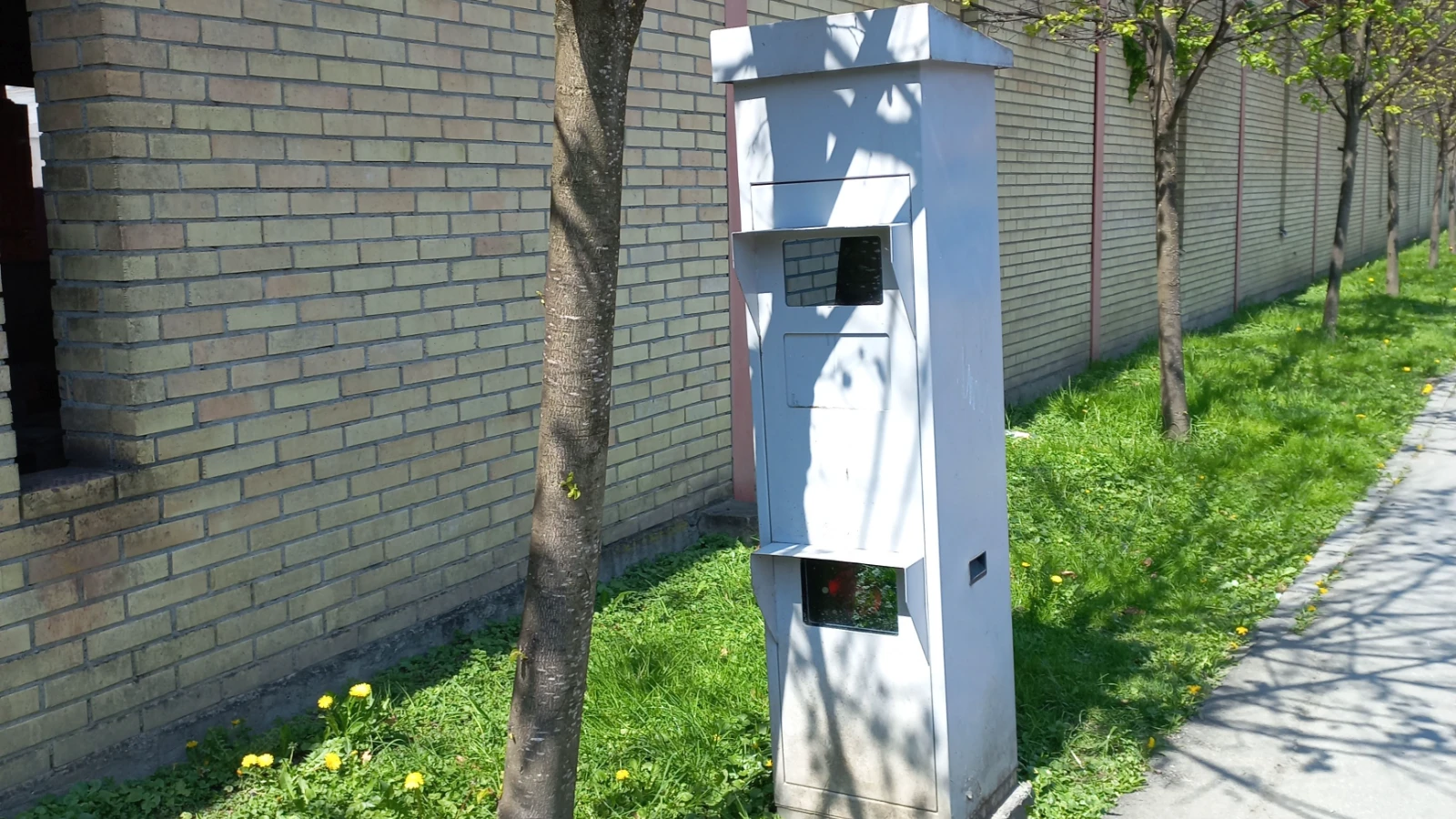 Hellgrauer Radarkasten auf einem Grünstreifen zwischen einem jungen Baum und einem Gehweg, dahinter eine lange Ziegelmauer