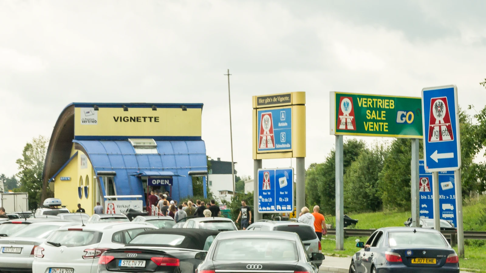 Autoschlange vor einem gelb-blauen Verkaufshäuschen mit der Aufschrift Vignette am Grenzübergang Suben, daneben die Schilder Hier gibt’s die Vignette und Vertrieb Sale Vente, vor dem Häuschen warten Menschen